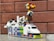 A colorful Lego model resembling a vehicle with a 'Police' label on the front. It has a mix of various bricks in colors like purple, orange, green, and blue, forming the base and body. On top, a multicolored structure with a wheel adds a playful detail. The background is a brown brick wall.