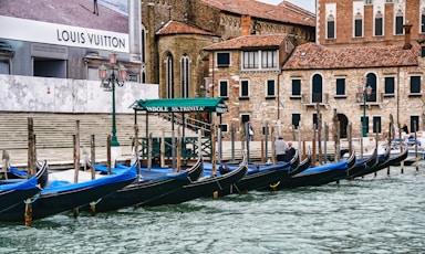 Several gondolas are moored at a canal in front of historical brick buildings with arched windows and tiled roofs. A large advertisement for a luxury brand is visible on a wall, and there's a green canopy labeled 'Gondole SS.Trinita'. The scene includes a few people walking along the water's edge and features classic lantern-style streetlights.
