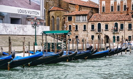 Several gondolas are moored at a canal in front of historical brick buildings with arched windows and tiled roofs. A large advertisement for a luxury brand is visible on a wall, and there's a green canopy labeled 'Gondole SS.Trinita'. The scene includes a few people walking along the water's edge and features classic lantern-style streetlights.