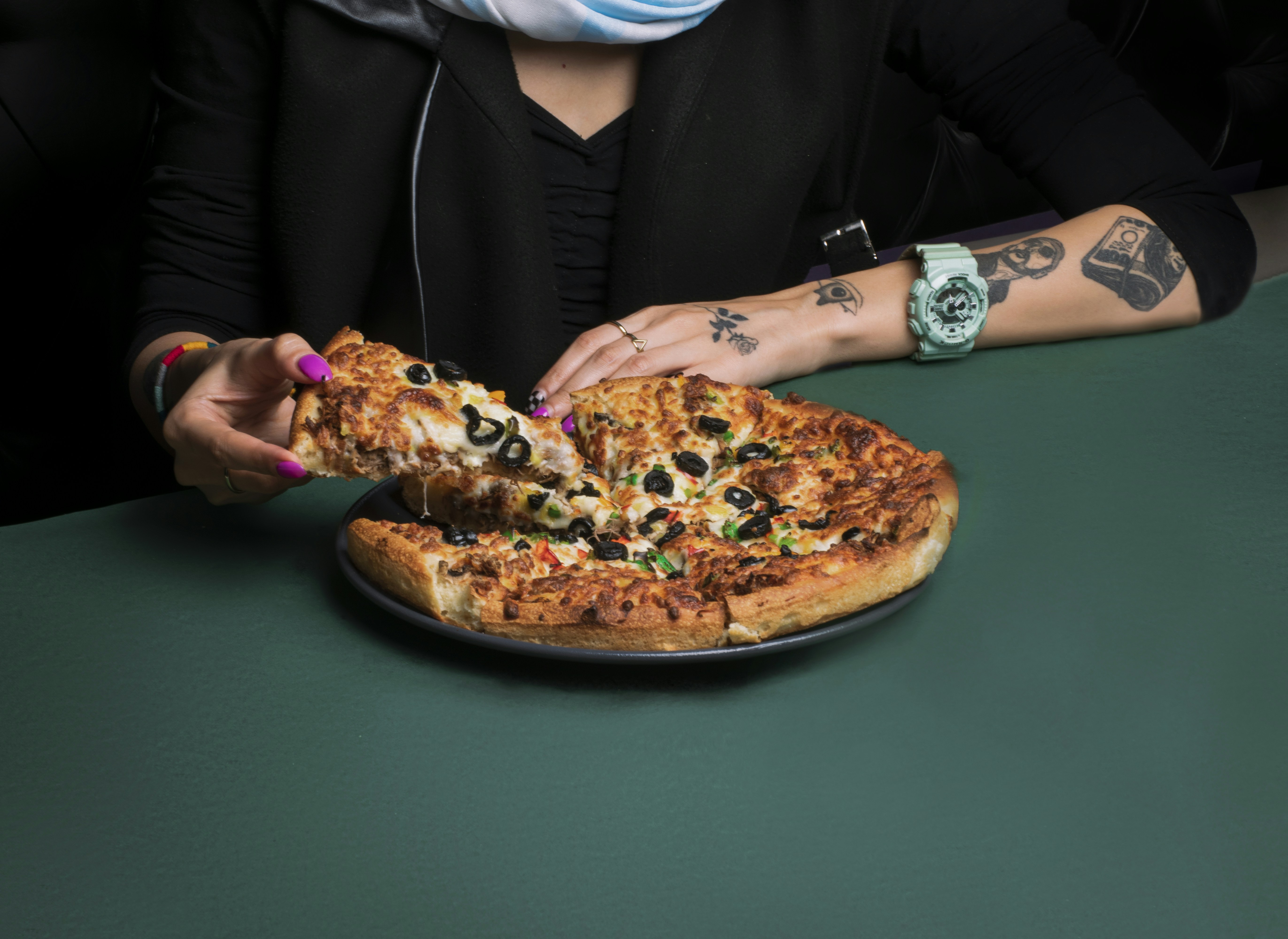A person is holding a slice of pizza with toppings including olives and green peppers, seated at a table. The individual has tattoos on their arm and wears a large, light-colored watch.