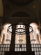 A grand, ornately designed interior space with large, circular chandeliers hanging from an arched ceiling. The architecture features tall arches and intricately patterned windows, allowing soft light to fill the area. Intricate designs and Arabic calligraphy adorn the upper sections of the walls.