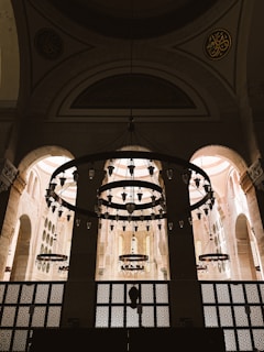 A grand, ornately designed interior space with large, circular chandeliers hanging from an arched ceiling. The architecture features tall arches and intricately patterned windows, allowing soft light to fill the area. Intricate designs and Arabic calligraphy adorn the upper sections of the walls.