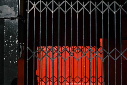 A metal gate with vertical bars casts shadows against a bright red wall, creating a geometric pattern. The black gate contrasts sharply with the intense red backdrop, adding depth and focus to the scene.