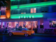 An art deco building with neon lights, featuring the name 'Avalon' in prominent green letters. People are casually dining or socializing outside under the awning. A vintage yellow and white car is parked in front of the establishment.