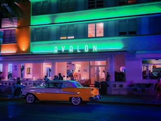 An art deco building with neon lights, featuring the name 'Avalon' in prominent green letters. People are casually dining or socializing outside under the awning. A vintage yellow and white car is parked in front of the establishment.