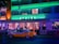 An art deco building with neon lights, featuring the name 'Avalon' in prominent green letters. People are casually dining or socializing outside under the awning. A vintage yellow and white car is parked in front of the establishment.