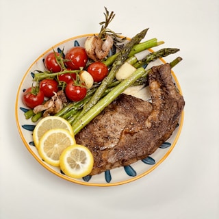 A colorful plate of food with a well-cooked steak, accompanied by asparagus, cherry tomatoes, roasted garlic, and slices of lemon.