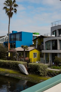 A vibrant residential area features modern houses painted in bright colors, including blue, yellow, and green. A palm tree stands tall to the left, and a kayak is placed upside down near the water along the edge of the canal. The area is surrounded by shrubs and greenery, with a clear sky overhead.