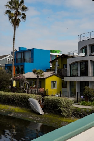 A vibrant residential area features modern houses painted in bright colors, including blue, yellow, and green. A palm tree stands tall to the left, and a kayak is placed upside down near the water along the edge of the canal. The area is surrounded by shrubs and greenery, with a clear sky overhead.