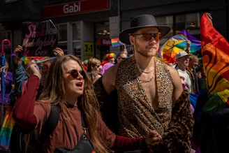A lively protest or parade featuring a diverse group of people. The scene includes vibrant rainbow flags and colorful clothing, with two central figures—one wearing a leopard print top and sunglasses, and another with long hair and glasses, appearing vocal and expressive. People are holding signs and flags with messages of pride and equality.