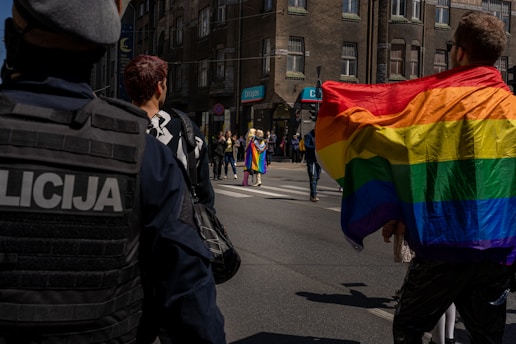 A street scene captures individuals participating in a parade or protest. One person prominently displays a large rainbow flag, a symbol commonly associated with LGBTQ+ pride. Several people can be seen wearing colorful clothing in the background, and at least one person appears to be a police officer monitoring the scene. The event occurs in an urban setting with brick buildings in the background.