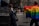 A street scene captures individuals participating in a parade or protest. One person prominently displays a large rainbow flag, a symbol commonly associated with LGBTQ+ pride. Several people can be seen wearing colorful clothing in the background, and at least one person appears to be a police officer monitoring the scene. The event occurs in an urban setting with brick buildings in the background.