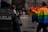 A street scene captures individuals participating in a parade or protest. One person prominently displays a large rainbow flag, a symbol commonly associated with LGBTQ+ pride. Several people can be seen wearing colorful clothing in the background, and at least one person appears to be a police officer monitoring the scene. The event occurs in an urban setting with brick buildings in the background.