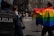 A street scene captures individuals participating in a parade or protest. One person prominently displays a large rainbow flag, a symbol commonly associated with LGBTQ+ pride. Several people can be seen wearing colorful clothing in the background, and at least one person appears to be a police officer monitoring the scene. The event occurs in an urban setting with brick buildings in the background.