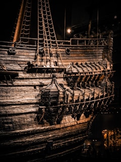 An intricate and well-preserved wooden ship with detailed carvings on its hull and a series of evenly spaced portholes. The ropes and masts are prominently visible, and light casts dramatic shadows on the weathered wood surface.
