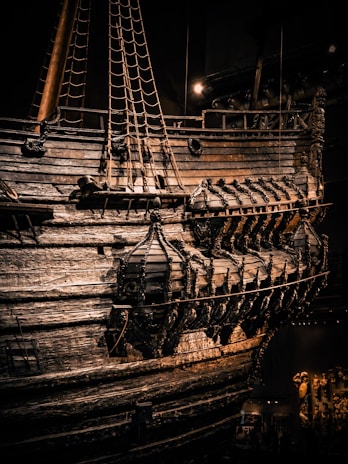 An intricate and well-preserved wooden ship with detailed carvings on its hull and a series of evenly spaced portholes. The ropes and masts are prominently visible, and light casts dramatic shadows on the weathered wood surface.