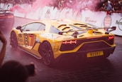A yellow sports car with black accents is driving on a paved road. It has a number 87 decal, Kappa and Fresh Scents logos, and a license plate reading B3 SVJ. There is pink smoke in the air and a crowd watching behind safety barriers.