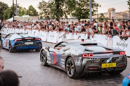 A crowd of people is gathered along a street, watching two sports cars that are decorated with various logos and designs. The cars are moving along a stone-paved road, surrounded by onlookers who are standing behind barriers. Trees and buildings can be seen in the background, suggesting an outdoor event or rally.