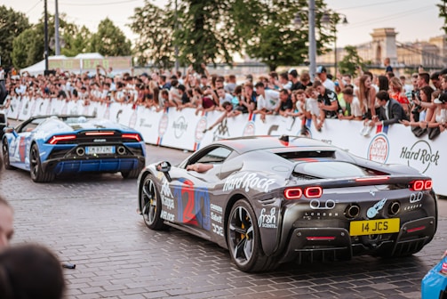 A crowd of people is gathered along a street, watching two sports cars that are decorated with various logos and designs. The cars are moving along a stone-paved road, surrounded by onlookers who are standing behind barriers. Trees and buildings can be seen in the background, suggesting an outdoor event or rally.
