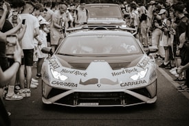 A sports car decorated with various logos is surrounded by a crowd of people on a street. The car is prominently displaying logos for brands like Hard Rock and Carrera. People around are taking photos and videos, capturing the moment. The scene seems to be part of an event or parade.