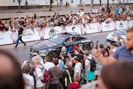A crowd of people stands along a barricade, excitedly watching and taking pictures as a black sports car with various logos and brand stickers drives past. The audience appears engaged, with several individuals holding up phones to capture the moment.