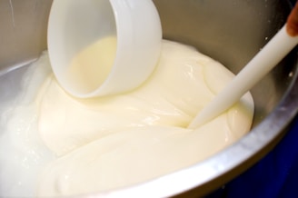 A thick, creamy white substance is being stirred in a large, metallic bowl with a white spatula. A white plastic container is partially submerged in the mixture.