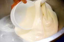 A person is pouring thick, creamy white liquid from one container into another stainless steel container.