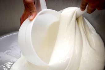 A close-up view of a white, creamy substance being poured and mixed with a large scoop in a smooth, flowing manner. The hands of a person are visible, holding the scoop and managing the thick texture.