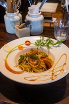 A plate of pasta garnished with herbs and vegetables, presented on a wooden table. The white plate is decorated with vibrant green herbs, sliced carrots, and an artful drizzle of sauce around the edges. In the background, there are ceramic jars holding various utensils, a glass of water, and a wooden organizer containing napkins and a smartphone.
