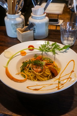 A plate of pasta garnished with herbs and vegetables, presented on a wooden table. The white plate is decorated with vibrant green herbs, sliced carrots, and an artful drizzle of sauce around the edges. In the background, there are ceramic jars holding various utensils, a glass of water, and a wooden organizer containing napkins and a smartphone.