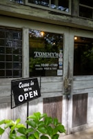 A rustic storefront with a wooden door and stained glass window. A sign indicates the shop is open, and there are lush green plants in the foreground. The shop's name is visible, along with its opening hours and days of closure.