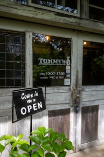 A rustic storefront with a wooden door and stained glass window. A sign indicates the shop is open, and there are lush green plants in the foreground. The shop's name is visible, along with its opening hours and days of closure.