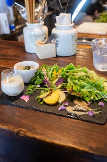 A wooden table with a black slate serving platter displaying a variety of food items, including fresh green salad leaves, sliced yellow potatoes, vibrant purple petals, and various sauces. Accompanying the dish is a glass with a thick beverage and a small white bowl containing a dark condiment. In the background, ceramic jars containing utensils like chopsticks, spoons, and paper napkins are visible, adding a casual dining feel.