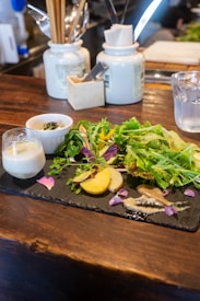 A wooden table with a black slate serving platter displaying a variety of food items, including fresh green salad leaves, sliced yellow potatoes, vibrant purple petals, and various sauces. Accompanying the dish is a glass with a thick beverage and a small white bowl containing a dark condiment. In the background, ceramic jars containing utensils like chopsticks, spoons, and paper napkins are visible, adding a casual dining feel.