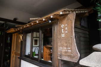 A small wooden storefront with a rustic aesthetic is adorned with a large wooden sign displaying the name 'etu' in both Latin and Japanese script. The building features a small overhang roof and a wooden framed window. Inside, there are objects like a plant and some dishware. The colors are earthy and warm, contributing to a cozy atmosphere.