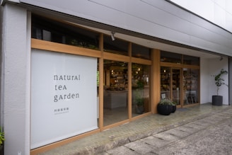 A modern storefront with large glass windows framed by light wood. The left side features a large sign with the text 'natural tea garden' in a minimalist font. The interior is visible through the windows, displaying shelves with various products. Potted plants are placed outside the entrance, contributing to a welcoming atmosphere.