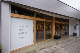 A modern storefront with large glass windows framed by light wood. The left side features a large sign with the text 'natural tea garden' in a minimalist font. The interior is visible through the windows, displaying shelves with various products. Potted plants are placed outside the entrance, contributing to a welcoming atmosphere.