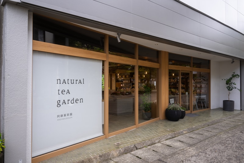 A modern storefront with large glass windows framed by light wood. The left side features a large sign with the text 'natural tea garden' in a minimalist font. The interior is visible through the windows, displaying shelves with various products. Potted plants are placed outside the entrance, contributing to a welcoming atmosphere.