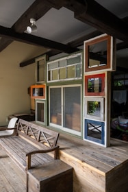 A cozy interior features a wooden bench and a wall composed of variously sized and colored window frames. The space is designed with rustic and eclectic elements, including exposed wooden beams on the ceiling and a mix of textures and finishes.