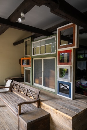 A cozy interior features a wooden bench and a wall composed of variously sized and colored window frames. The space is designed with rustic and eclectic elements, including exposed wooden beams on the ceiling and a mix of textures and finishes.