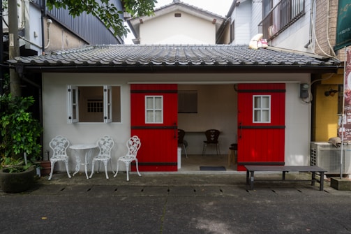 A small house with striking red doors and a tiled roof, flanked by white window frames. In front, there's a set of white ornate metal chairs and a table on a stone pavement. A few potted plants add greenery to the scene, while a minimalistic interior is visible through the open door.