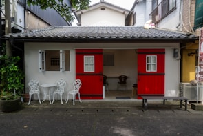 A small house with striking red doors and a tiled roof, flanked by white window frames. In front, there's a set of white ornate metal chairs and a table on a stone pavement. A few potted plants add greenery to the scene, while a minimalistic interior is visible through the open door.