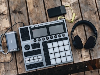 An electronic music production controller with multiple pads and knobs lies on a rustic wooden table. Beside it are a pair of over-ear headphones and a small, portable battery pack with a coiled yellow cable.