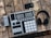 An electronic music production controller with multiple pads and knobs lies on a rustic wooden table. Beside it are a pair of over-ear headphones and a small, portable battery pack with a coiled yellow cable.