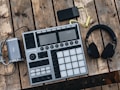 An electronic music production controller with multiple pads and knobs lies on a rustic wooden table. Beside it are a pair of over-ear headphones and a small, portable battery pack with a coiled yellow cable.