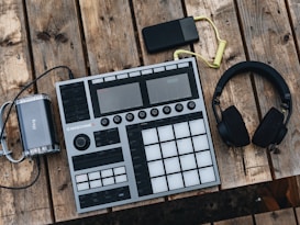 An electronic music production controller with multiple pads and knobs lies on a rustic wooden table. Beside it are a pair of over-ear headphones and a small, portable battery pack with a coiled yellow cable.