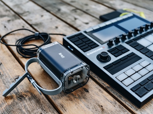 A music production device with various knobs, pads, and buttons sits on a rustic wooden surface. Next to it is a portable power bank with a cable coiled beside it.
