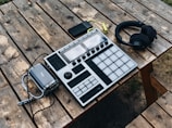 A wooden picnic table holds electronic music equipment, including a music production controller with pads and knobs, a set of black headphones, a portable battery pack, and a smartphone. The arrangement suggests an outdoor setting for music creation.