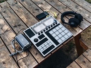 A wooden picnic table holds electronic music equipment, including a music production controller with pads and knobs, a set of black headphones, a portable battery pack, and a smartphone. The arrangement suggests an outdoor setting for music creation.