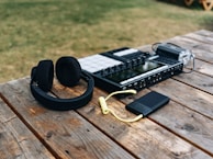 A pair of black headphones, a music production controller, a black portable electronic device with a yellow cable, and a handheld video recorder are placed on a wooden table outdoors. The scene features grassy surroundings.